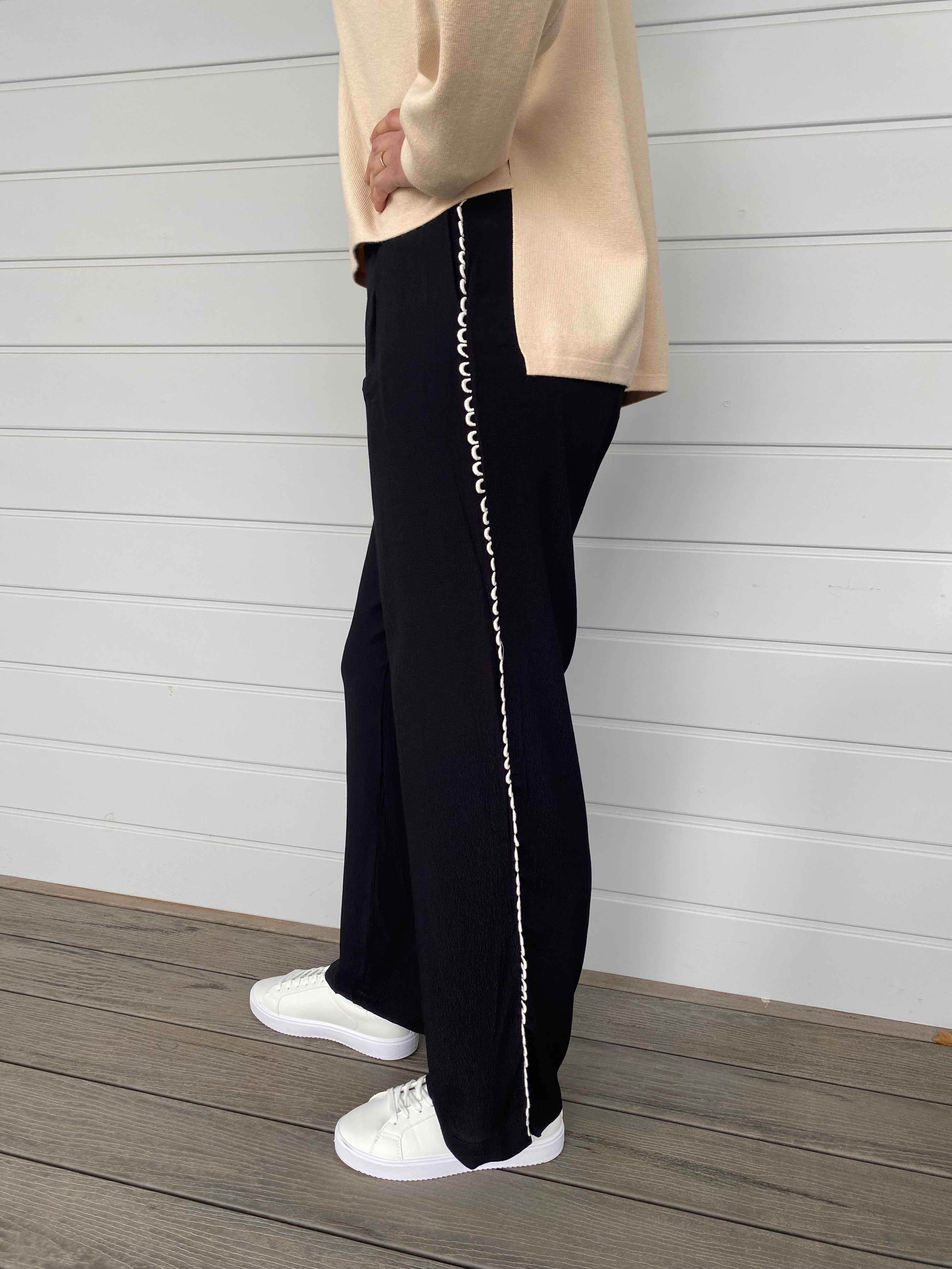 A woman dressed in a beige sweater and black pants stands confidently, showcasing a casual yet stylish outfit.