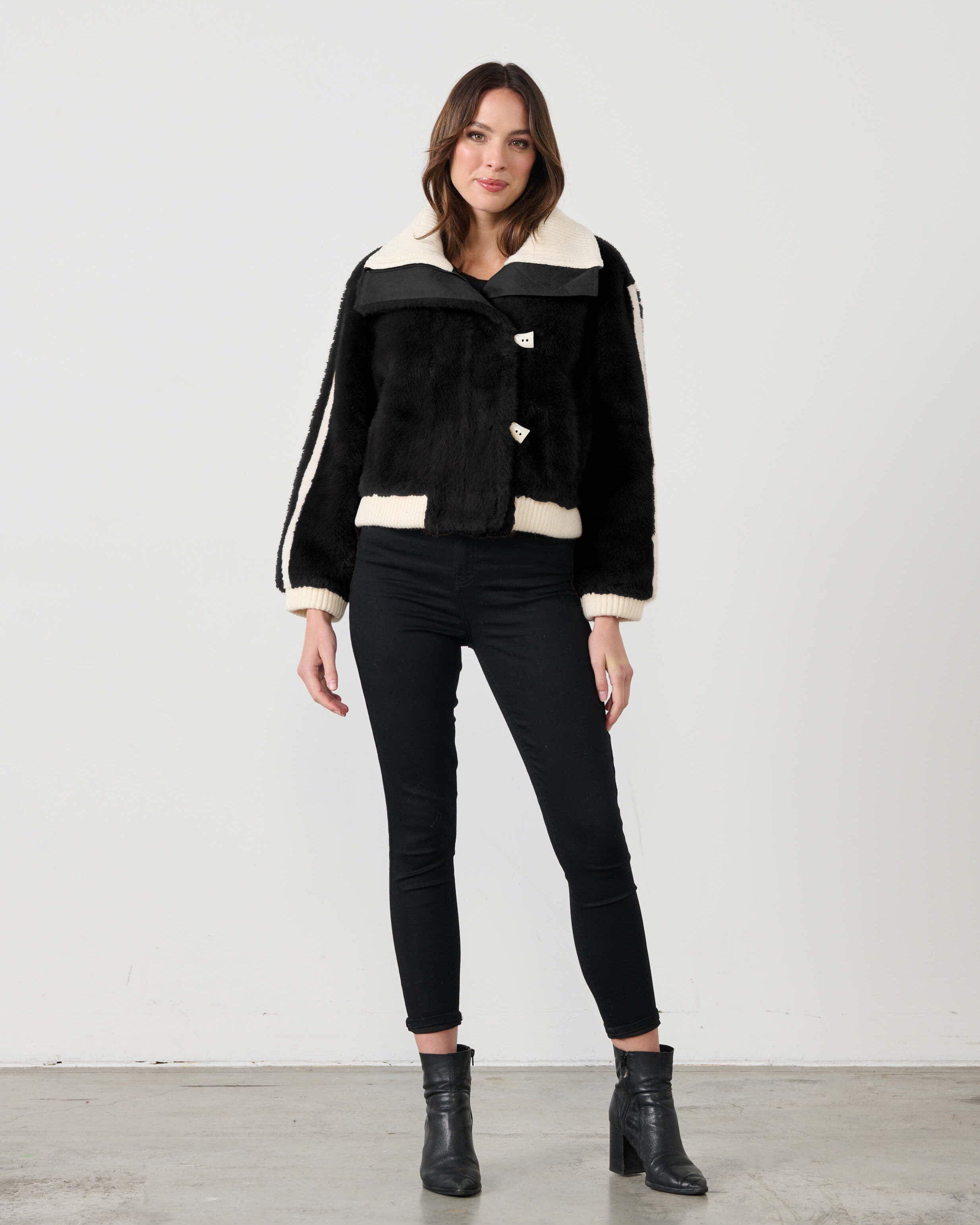 A woman is standing, wearing a black faux fur bomber jacket with white collar and zipper details, paired with black leggings and black boots.