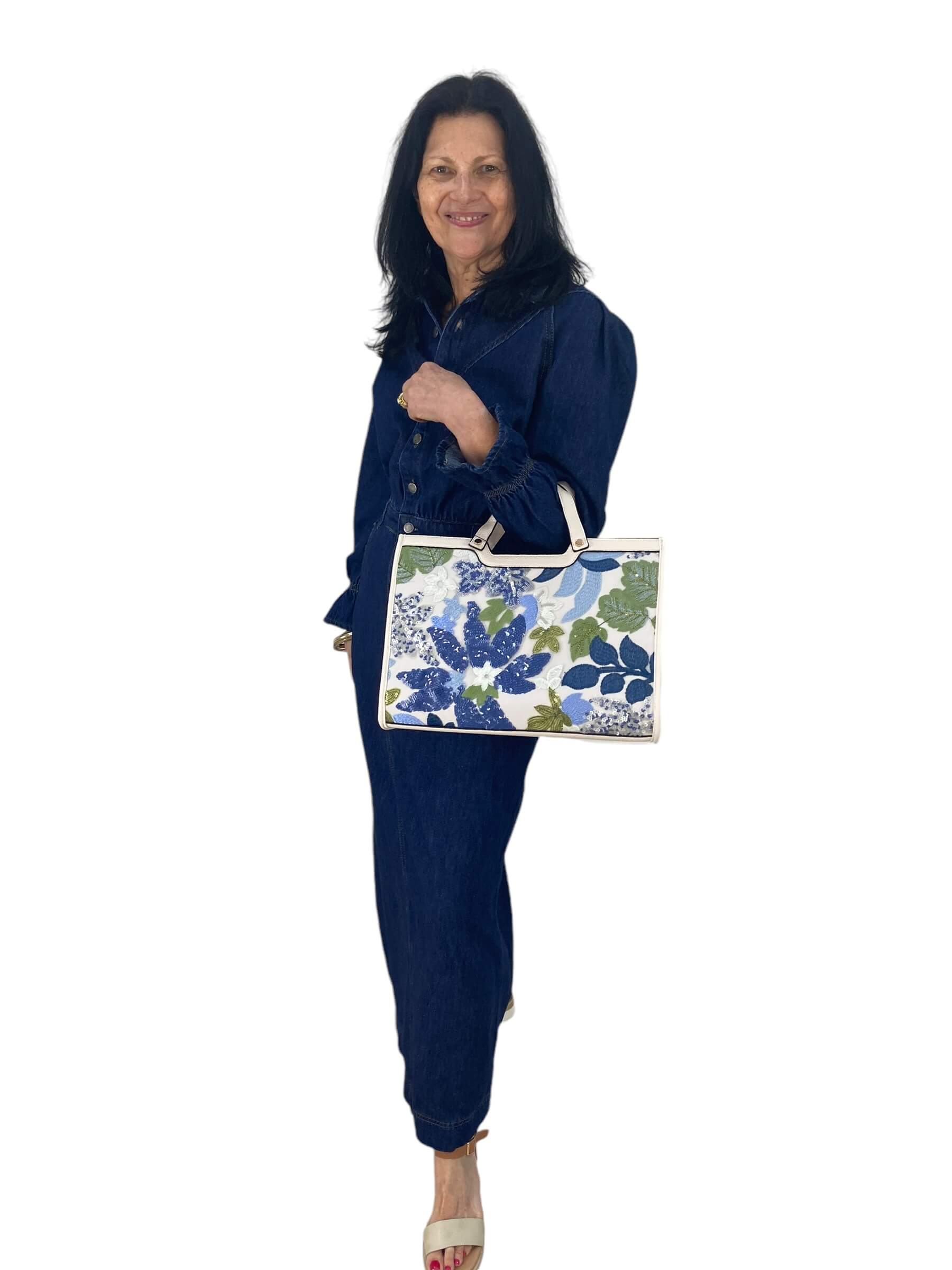 A woman in a blue dress smiles while holding a colorful floral bag, standing against a bright background.