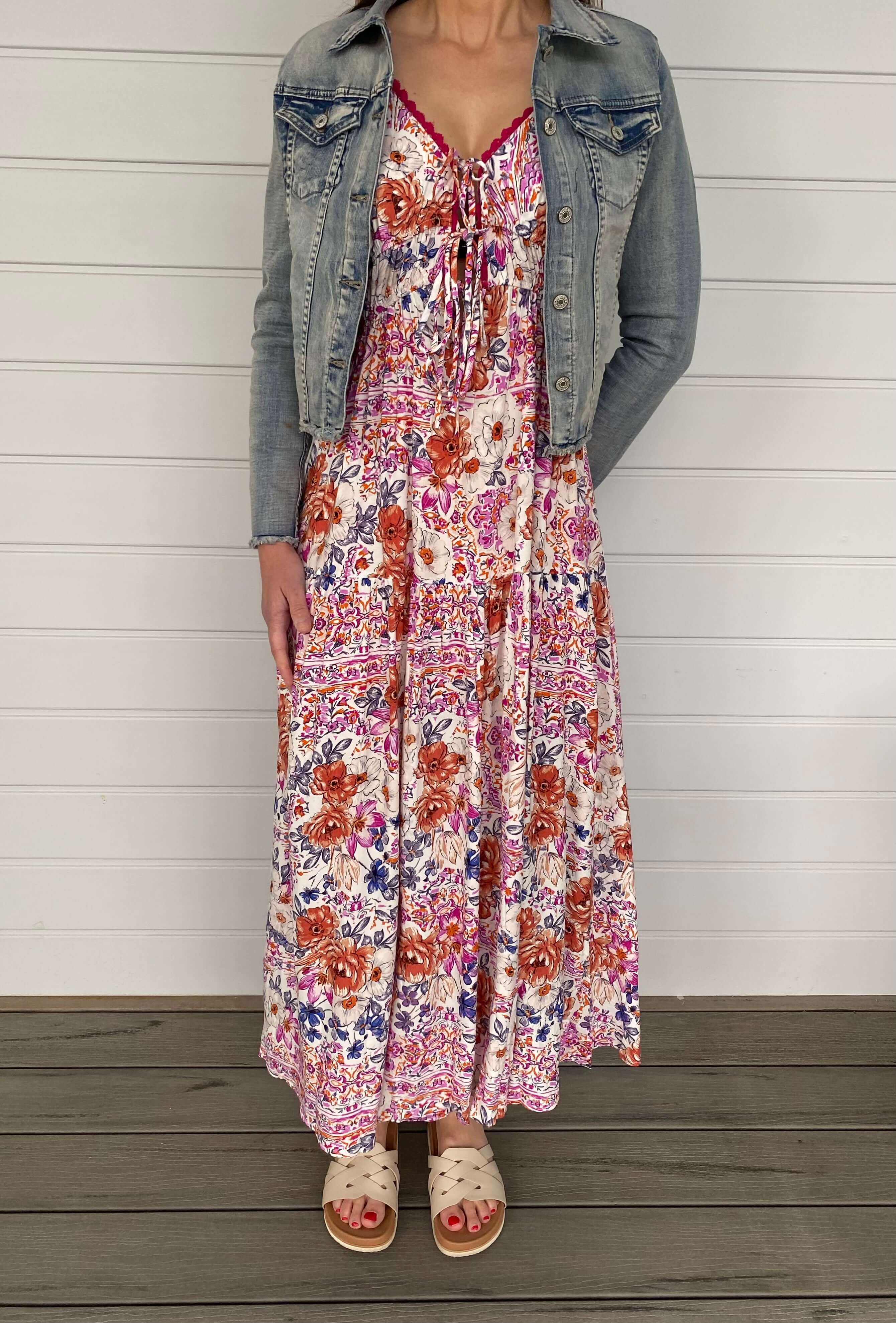 A woman wearing a floral dress and denim jacket stands happily, embodying a fresh and fashionable look in a sunny environment.
