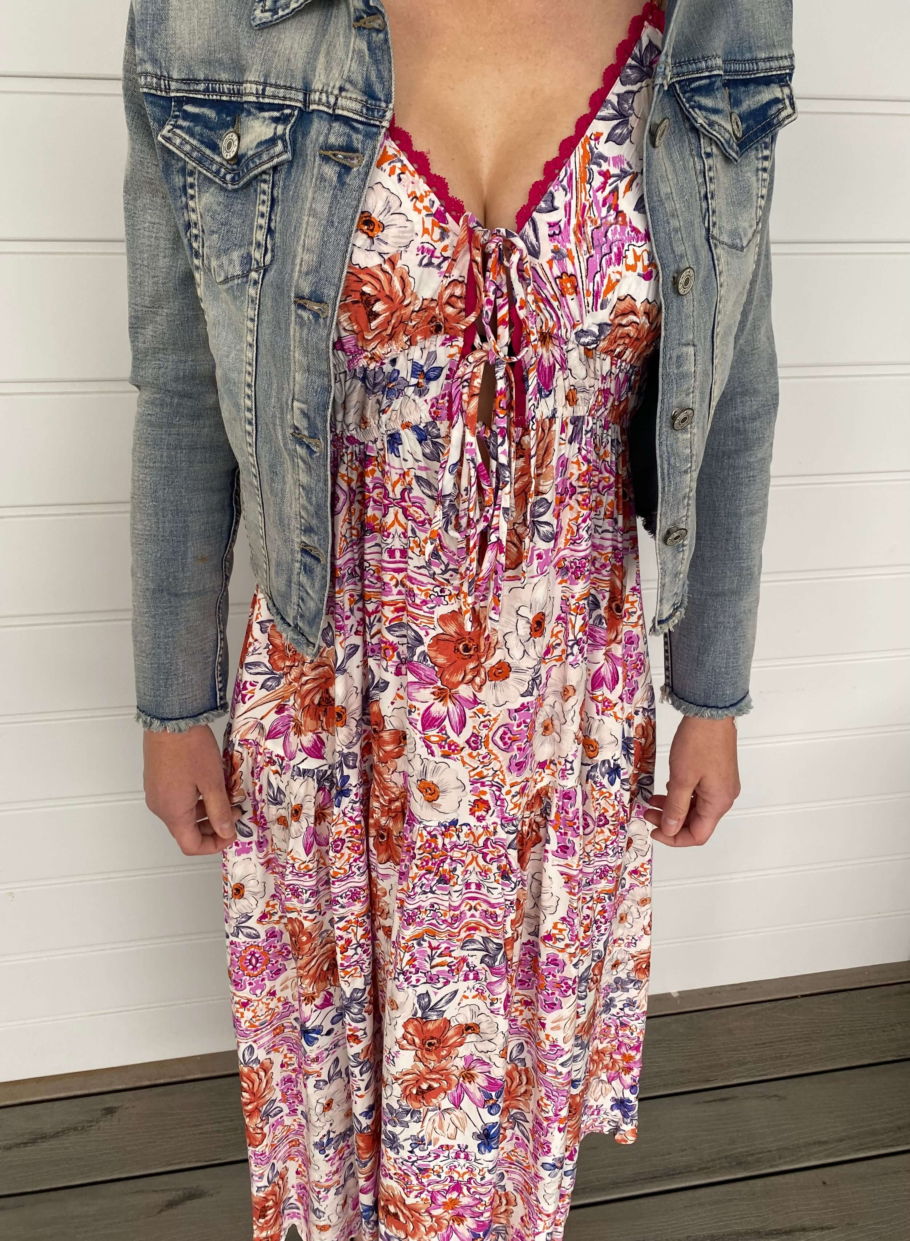 A woman wearing a floral dress and denim jacket stands happily, embodying a fresh and fashionable look in a sunny environment.