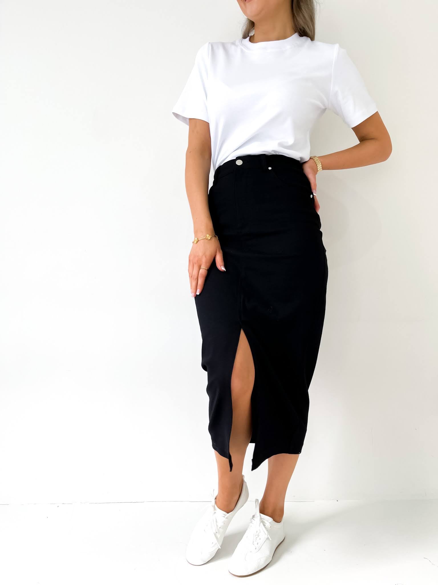 Woman standing in a black denim skirt and white top with a white background and white sneakers