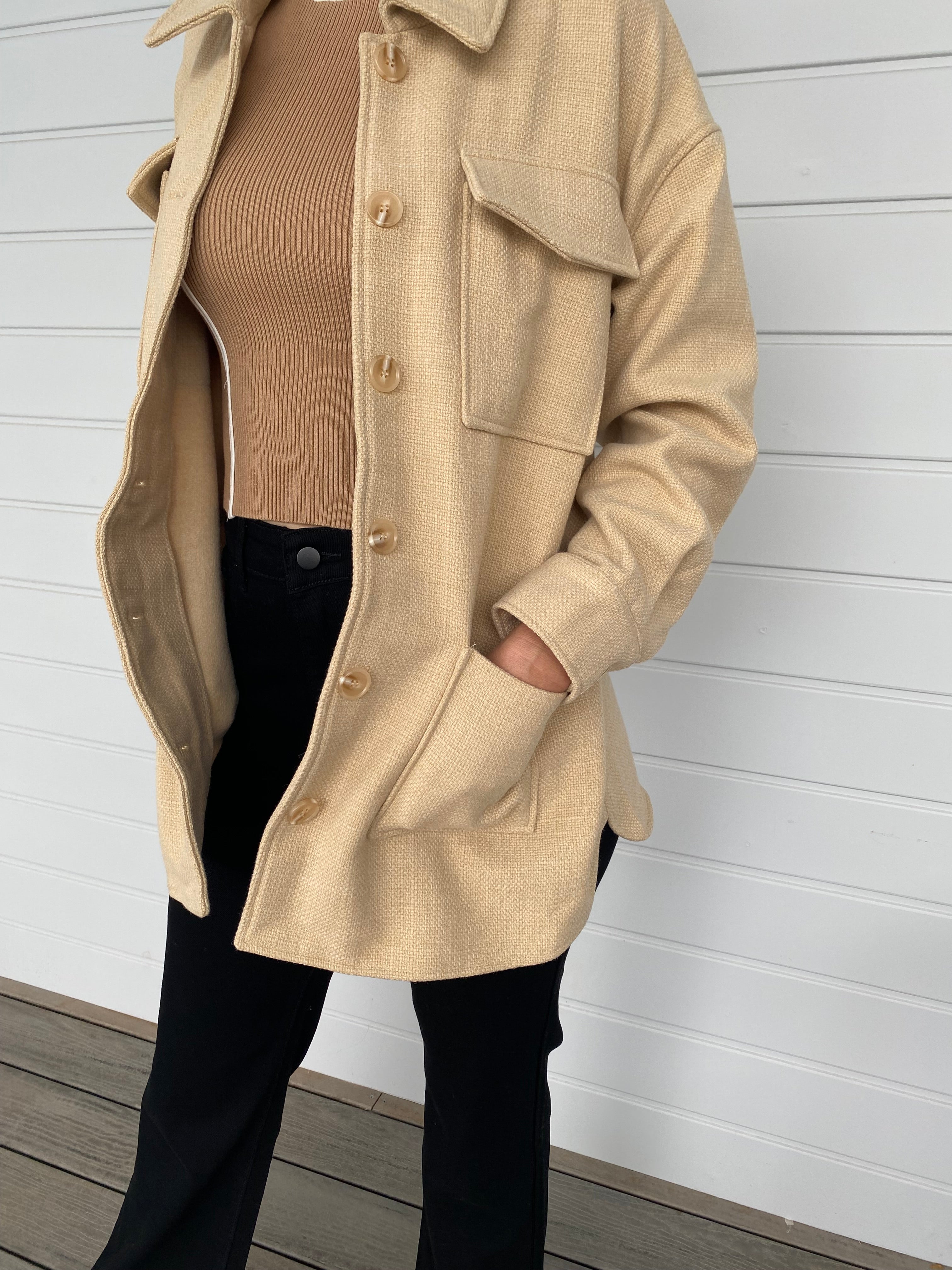 Woman standing in front of a white wall in a beige weed jacket and black jeans