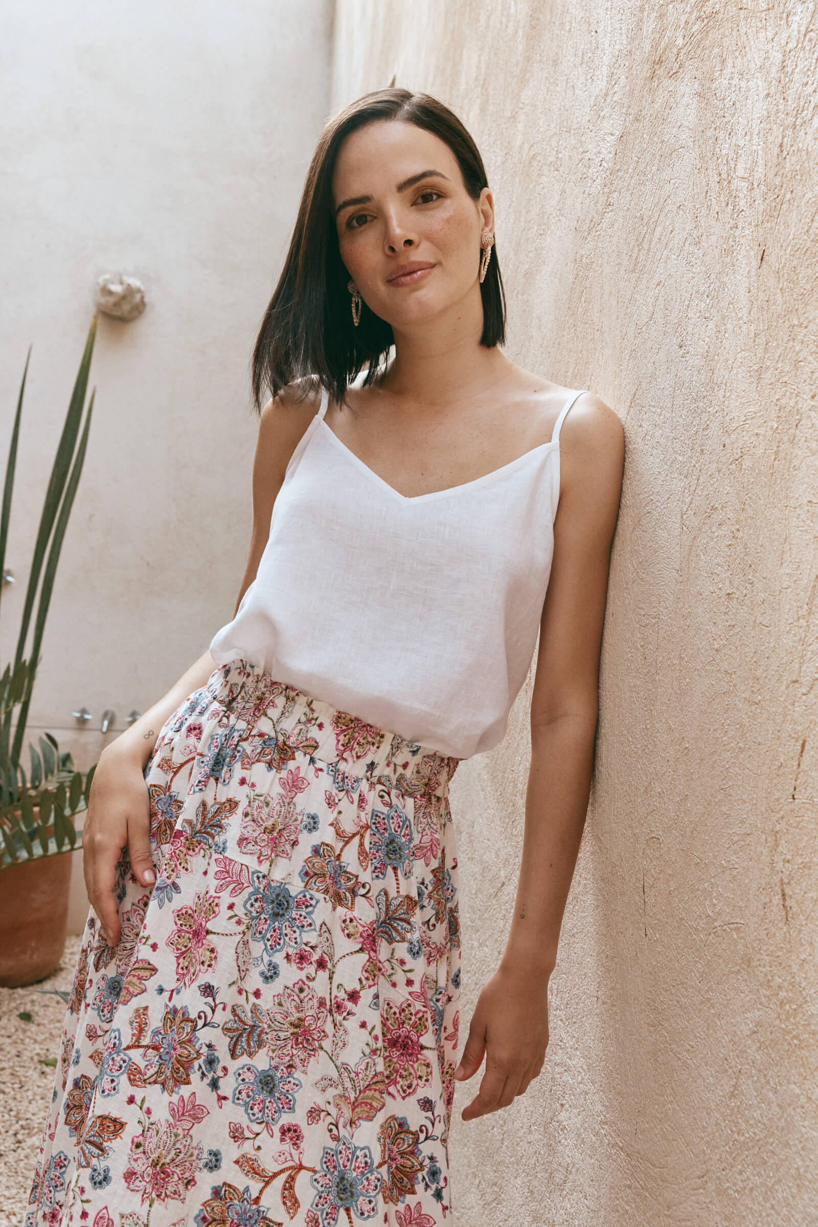 A woman in a white top and floral skirt leans casually against a wall, exuding a relaxed and stylish vibe.