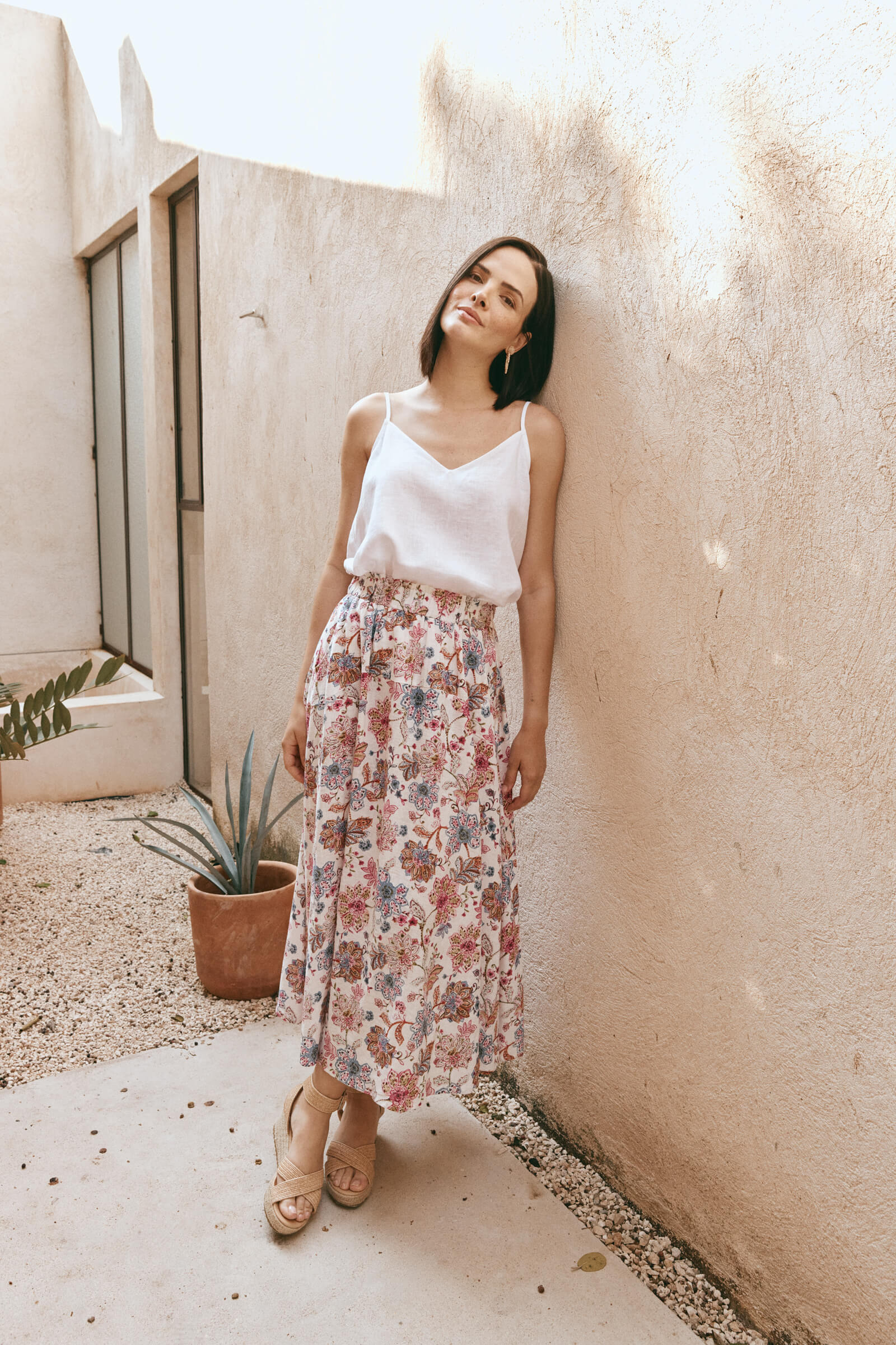 A woman in a white top and floral skirt leans casually against a wall, exuding a relaxed and stylish vibe.