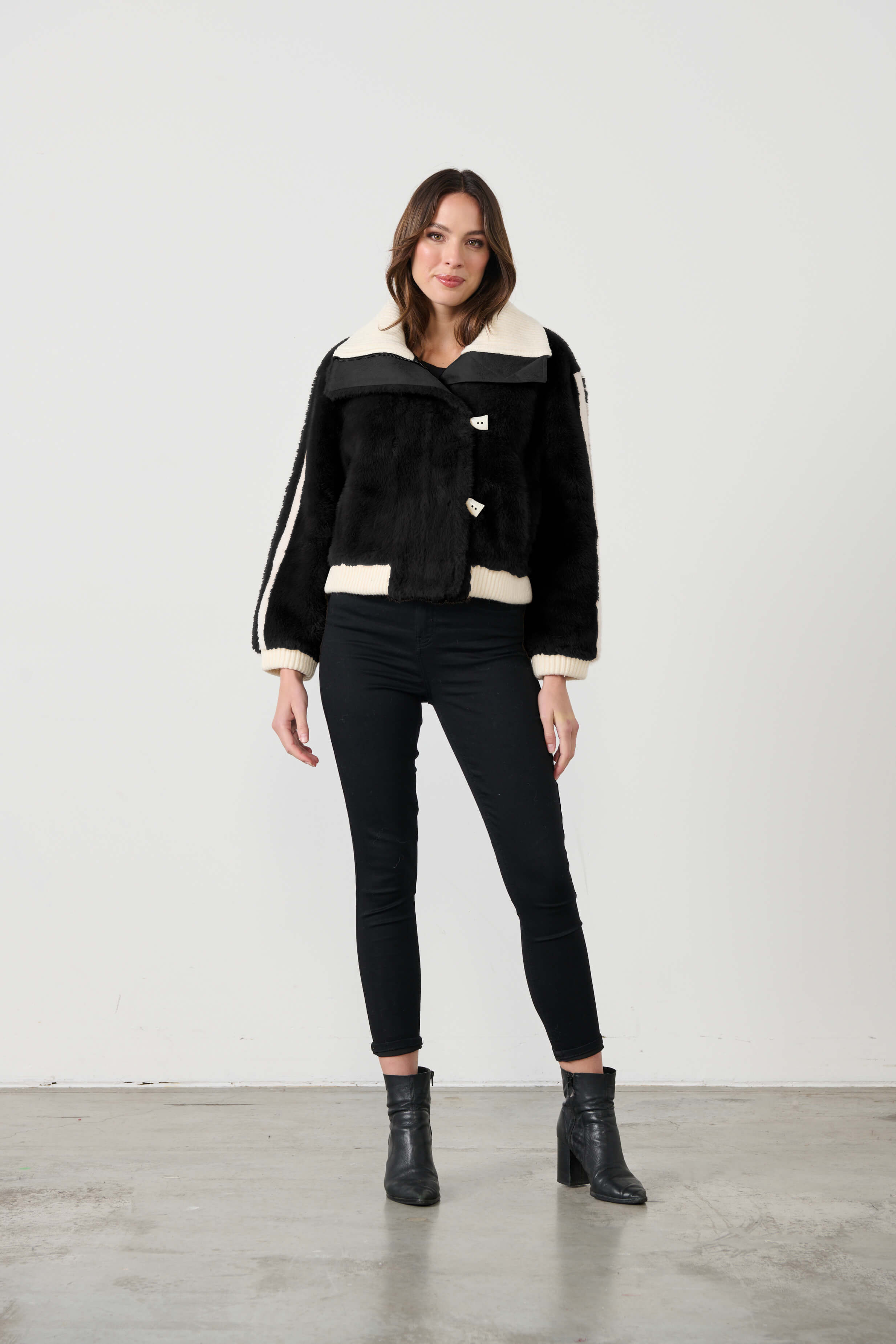 A woman is standing, wearing a black faux fur bomber jacket with white collar and zipper details, paired with black leggings and black boots.