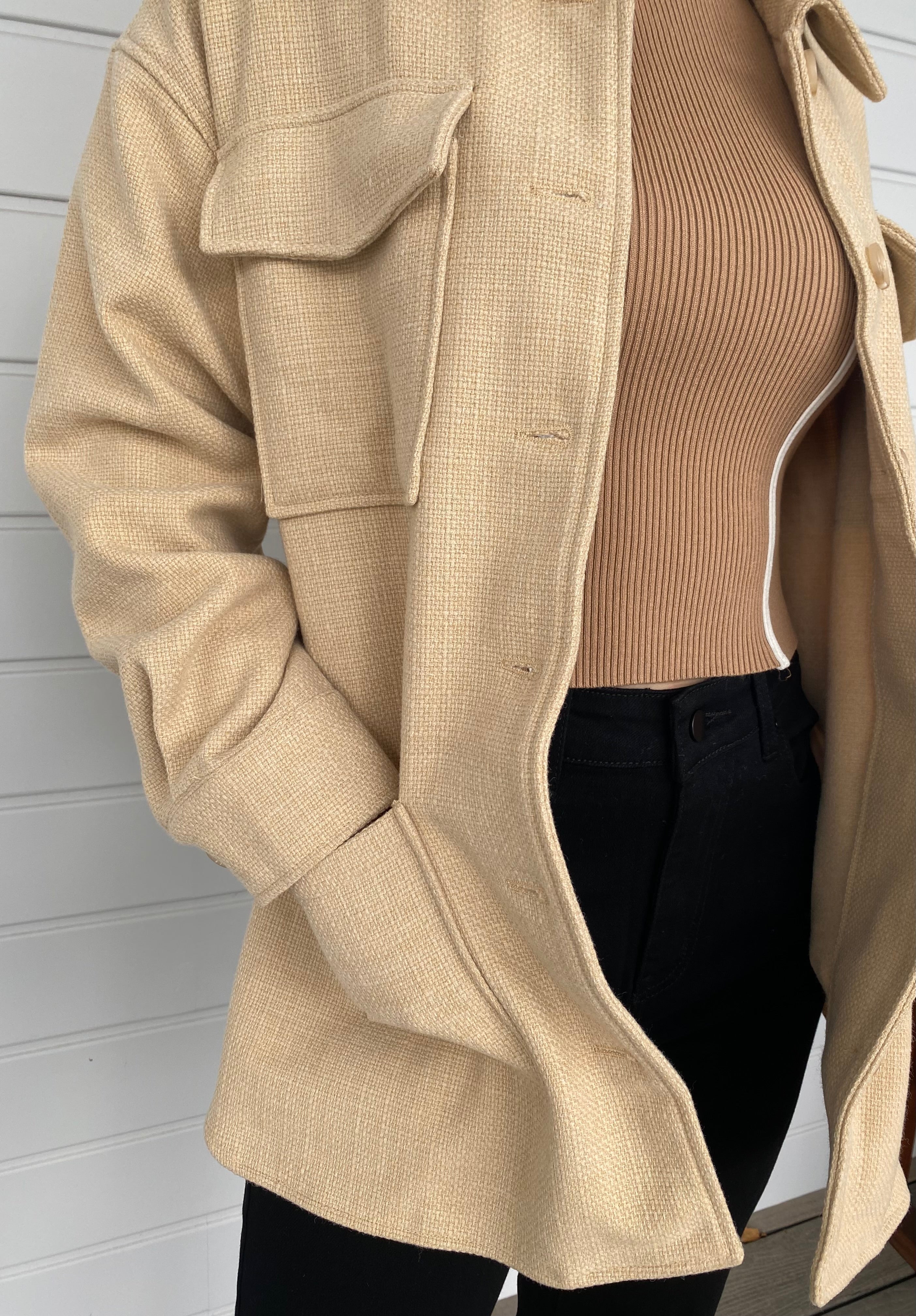 Woman standing in front of a white wall in a beige weed jacket and black jeans