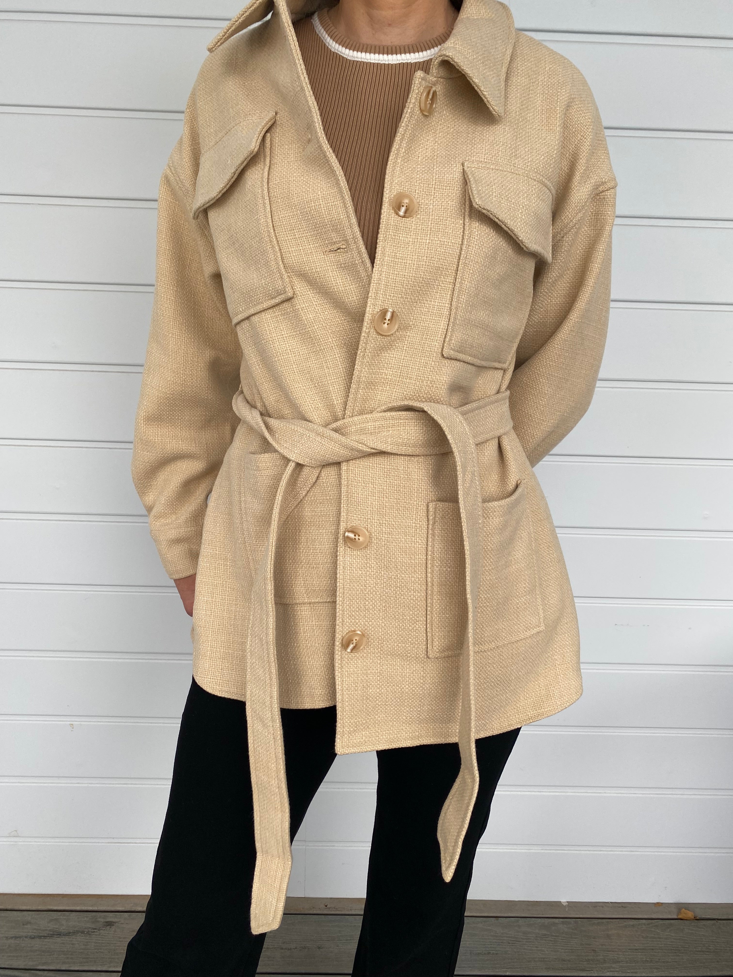 Woman standing in front of a white wall in a beige weed jacket and black jeans