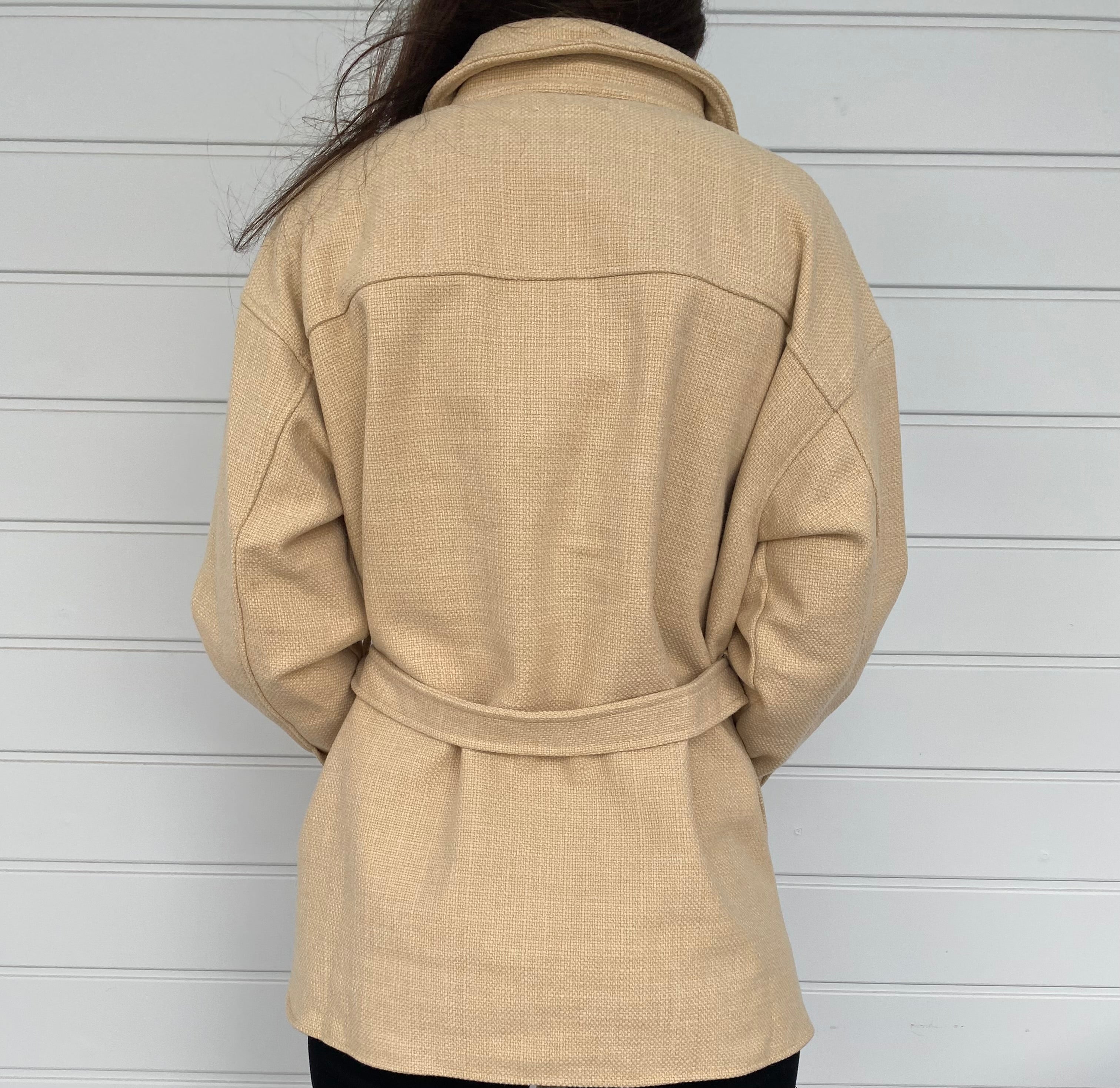 Woman standing in front of a white wall in a beige weed jacket and black jeans
