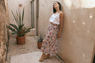  A woman in a white top and floral skirt leans casually against a wall, exuding a relaxed and stylish vibe.