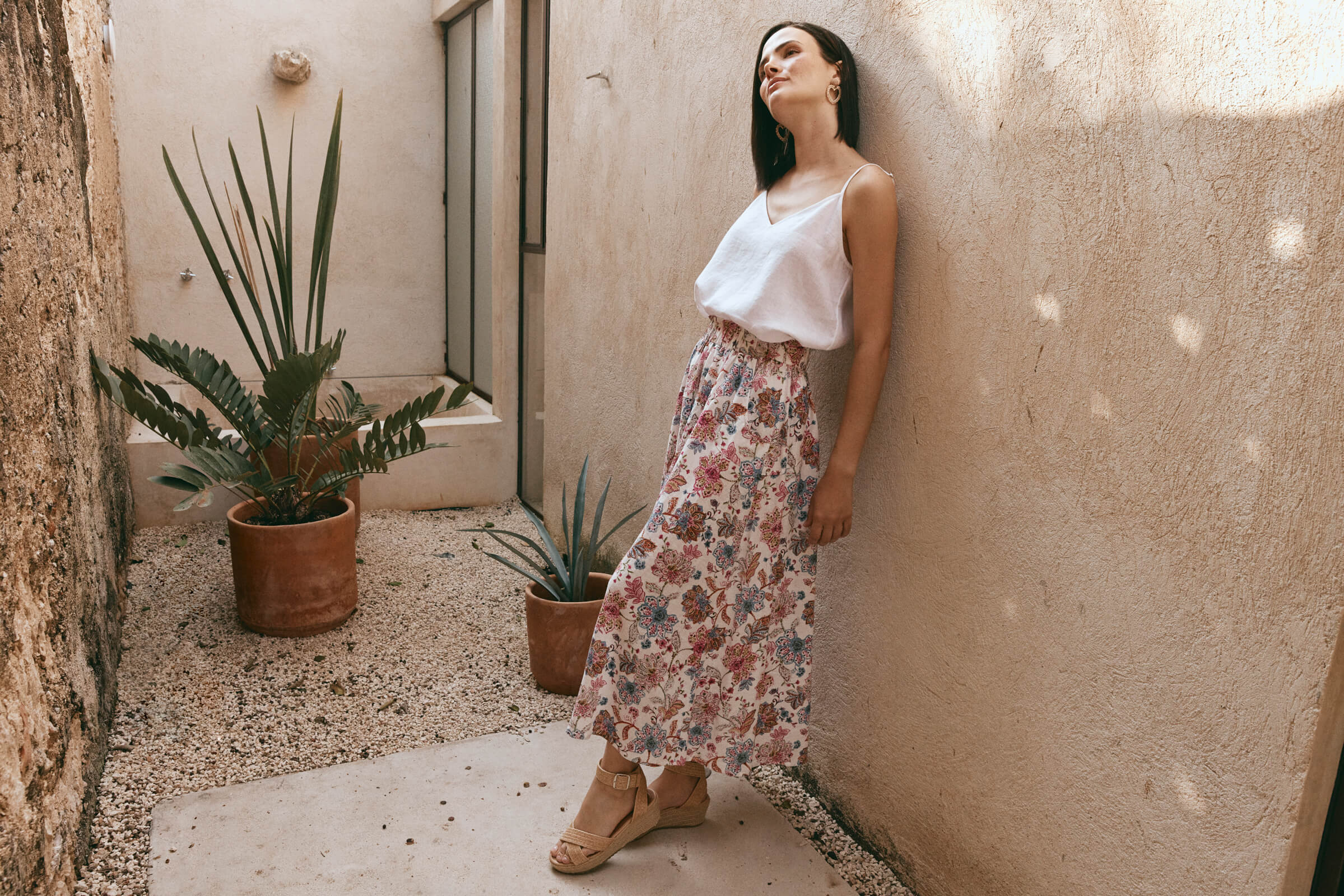  A woman in a white top and floral skirt leans casually against a wall, exuding a relaxed and stylish vibe.